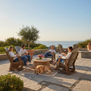 Five people relax on Kanyon Living Adirondack Poly Glider Swivel Chairs in Light Grey & Weathered Wood around a coffee table on a sunny patio with ocean views, as a golden retriever lounges near potted plants and eco-friendly furniture.