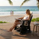 A woman relaxes in a Kanyon Living Adirondack Poly Glider Swivel Chair in Seashell & Black on the deck, reading by a small table with a coffee cup. Palm trees frame the view as a couple strolls along the shoreline in the background.