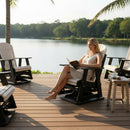 A woman sits barefoot on a wooden deck by a lake, reading in a Kanyon Living Adirondack Poly Glider Swivel Chair (Seashell & Black), surrounded by palm trees, empty outdoor poly lumber furniture, calm water, and green trees.
