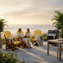 A couple relaxes on Kanyon Living Adirondack Poly Glider Swivel Chairs in Yellow & Cedar by the beach at sunset, with a grill, table with wine glasses, lush plants, and a scenic ocean view in the background.