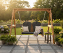 The Kanyon Living Poly Lumber 4′ Adirondack Swing in Black & Deluxe Antique Mahogany sits on a stone patio with two chairs and white cushions, a blanket draped over the swing, and nearby potted plants. Green lawn and trees are in the background.