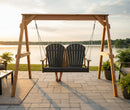 The Kanyon Living Poly Lumber 4′ Adirondack Swing in Black & Deluxe Antique Mahogany hangs on a patio by a tranquil lake at sunset, with someone relaxing on it among lush plants and cozy outdoor furnishings.