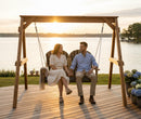 A couple enjoys the sunset on a lakeside deck, holding hands and smiling as they relax in the Kanyon Living Poly Lumber 4′ Adirondack Swing Black & Deluxe Antique Mahogany, surrounded by peaceful water, flowers, and greenery.