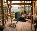 A woman sits on the Kanyon Living Poly Lumber 4′ Adirondack Swing in Dark Green and Cedar Wood, holding a mug and smiling as her dog relaxes. The rustic porch with flowers offers beautiful sunset views of trees and distant mountains.