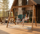 A woman sits on a Kanyon Living Poly Lumber 4′ Adirondack Swing in Dark Green and Cherry Wood, holding a mug and smiling on a log cabin deck. A blanket, pillows, outdoor chairs, and a fire pit are in the background.