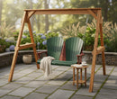The Kanyon Living Poly Lumber 4′ Adirondack Swing in dark green and cherry wood sits on a stone patio, draped with a white blanket. A side table holds coffee, books, and flowers amid lush plants and hydrangeas for an inviting outdoor scene.