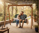 A couple relaxes on the Kanyon Living Poly Lumber 4′ Adirondack Swing in dark green and cherry wood under a pergola, surrounded by flowers and plants, enjoying a peaceful sunny day on quality outdoor furniture.