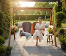 A woman in a light dress relaxes barefoot on the Kanyon Living Poly Lumber 4′ Adirondack Swing in Dark Wood and Orange, holding a cup. A small table with a drink and blanket sit nearby amid lush greenery, creating a tranquil outdoor setting.