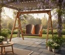 The Kanyon Living Poly Lumber 4′ Adirondack Swing in dark wood and orange sits under a pergola draped with blooming wisteria, surrounded by potted plants on a sunny stone patio. A round wooden table and more outdoor furniture are nearby.