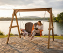 A woman relaxes barefoot on the Kanyon Living Poly Lumber 4′ Adirondack Swing in dark wood and orange, reading a book by the lake. A blanket and mug are beside her, with water, hills, and trees under a cloudy sky.