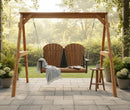 The Kanyon Living Poly Lumber 4′ Adirondack Swing Deluxe in Antique Mahogany and Black hangs from a wooden frame with two seats, paired with a small round table and coffee mug, surrounded by lush potted plants in a sunlit garden.