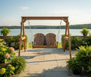 The Kanyon Living Poly Lumber 4′ Adirondack Swing Deluxe in Antique Mahogany and Black hangs on a stone patio overlooking a calm lake, surrounded by lush greenery and blooms—deluxe outdoor seating from Kanyon Living at its finest.