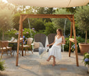 A woman in a white dress sits on the Kanyon Living Poly Lumber 4′ Adirondack Swing Deluxe Driftwood Grey and White, surrounded by garden chairs, potted plants, flowers, a tea table, and a sun shade on a sunny patio.