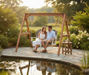 A couple relaxes on the Kanyon Living Poly Lumber 4′ Adirondack Swing Deluxe in Driftwood Grey and White, enjoying a sunny day by a pond surrounded by lush greenery and blooming flowers.
