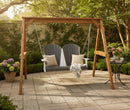 The Kanyon Living Poly Lumber 4′ Adirondack Swing Deluxe in Driftwood Grey and White sits on a stone patio, accented with beige pillows, a striped throw, flowers, greenery, and a tea set while sunlight streams through the trees.
