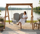 A woman in a white dress sits on a Kanyon Living Poly Lumber 4′ Adirondack Swing in light grey and weathered wood by a lake, surrounded by Adirondack chairs, outdoor patio furniture, potted lavender, and coffee cups on the stone patio.