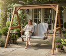 A woman smiles on the Kanyon Living Poly Lumber 4′ Adirondack Swing in light grey and weathered wood, surrounded by lush plants. Next to her, outdoor furniture includes a small table with cups and a book. Glass doors lead to the house in the background.
