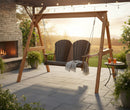 The Kanyon Living Poly Lumber 4′ Adirondack Swing Deluxe in Brazilian Walnut and Weathered Wood sits on a stone patio near a fireplace. String lights glow above, plants surround the area, two drinks rest nearby, and a blanket is draped over the swing at sunset.