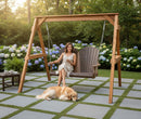 A woman in a light dress relaxes on the Kanyon Living Poly Lumber 4′ Adirondack Swing Deluxe in Brazilian Walnut and Weathered Wood, smiling with a drink as her golden retriever lounges nearby amid hydrangeas and trees.