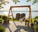 The Kanyon Living Poly Lumber 4′ Adirondack Swing Deluxe in Brazilian Walnut and Weathered Wood seats two on a stone patio by a lake, with outdoor furniture, a small table holding a drink and book, all surrounded by lush plants under a clear sky.