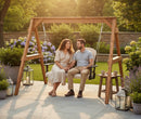 A couple relaxes on the Kanyon Living Poly Lumber 4′ Adirondack Swing in Seashell and Black, holding hands and smiling amidst blooming flowers. Lanterns, coffee cups, and a small table complete this cozy Kanyon Living porch scene.