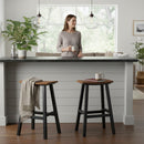 A woman with a mug stands behind a kitchen island, with two Kanyon Living Poly Lumber Saddle Stool Bar Height Deluxe Antique Mahogany and Black stools in front—one draped with a blanket—amid plants and pendant lights.