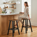 A woman in light clothing sits on a Kanyon Living Poly Lumber Saddle Stool Bar Height Deluxe in Antique Mahogany and Black at a kitchen island, holding a mug and smiling. Wooden cabinets, shelves, and natural light complete the space.