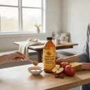 A bottle of Guenther Apiary Apple Cider Vinegar with Honey & “Mother” sits on a table with apples, bread, a honey bowl, and a honey dipper as two people reach in, one dipping honey, in a bright kitchen by the window.