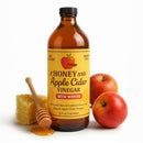 A bottle of Guenther Apiary Apple Cider Vinegar with Honey & “Mother” stands beside a honey dipper coated with honey, raw unfiltered honeycomb, and two red apples as honey drips onto the white surface.