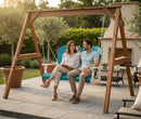 A smiling couple relaxes on a Kanyon Living Poly Lumber 4′ Adirondack Swing in Aruba Blue & White under a wooden frame, surrounded by eco-friendly outdoor furniture, potted plants, and seating on a sunny backyard patio.