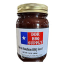A glass jar with a gold lid labeled DDR BBQ Supply Bacon Bourbon BBQ Sauce – 16 oz features a Texas flag design, address details, and highlights this smoky barbecue sauce’s bold Southern flavor.