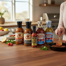 Five bottles from Bear & Burton's Ultimate Sauce Bundle are arranged on a kitchen counter with fresh vegetables, herbs, and a lemon, while someone stirs spices in a skillet as sunlight streams in through the window.