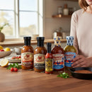 In a kitchen, a person stands by Bear & Burton's Ultimate Sauce Bundle—five versatile sauces—arranged on a wooden counter with herbs, sliced lemon, and cherry tomatoes.