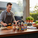 A smiling man in an apron prepares food outdoors, holding a fork with food. Bear & Burton's Ultimate Sauce Bundle—four versatile sauces—are on the counter, with fresh vegetables and herbs in the background.