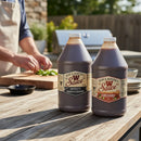 Two 64 oz bottles of Bear & Burton's W Sauce sit on an outdoor table near someone chopping herbs, ready to be used as the ultimate grilling marinade or in bulk for BBQ.