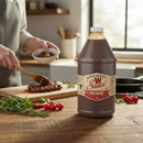 A 2-pack of Bear & Burton's W Sauce 64 oz Bulk Worcestershire sits on a wooden counter as someone preps food with fresh herbs and vegetables in the background, highlighting its use as a bold marinade or BBQ sauce.