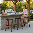 A woman sits on a Kanyon Living Poly Lumber Saddle Stool Bar Height Black and Deluxe Antique Mahogany by an outdoor kitchen island, holding a drink. A man stands by with food. The garden features flowers, greenery, weather-resistant furniture, and a grill.