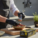 Wearing a gray apron and Crawford Black Mamba 3XL Black Food Prep Gloves, a person seasons raw meat chunks with pepper. A box of Crawford Black Mamba gloves sits beside a cutting board and potted herb plant on the counter.