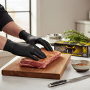 Wearing Crawford Black Mamba 3XL Black Food Prep Gloves (100 count), a person seasons raw meat on a wooden cutting board, with herbs, spices, a knife, and heavy-duty BBQ gloves also visible on the kitchen counter.