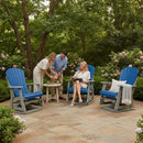 Four adults gather on a stone patio with lush greenery. Two women relax in blue and light grey Kanyon Living Adirondack Poly Glider Swivel Chairs, while another woman and a man prepare drinks at a small round table.
