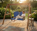 A woman relaxes on the Kanyon Living Poly Lumber 4′ Adirondack Swing in blue and light grey, reading a book in a sunlit garden. A small table with a mug is beside her on the stone patio surrounded by blooming hydrangeas.