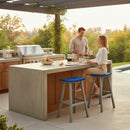 A man and woman enjoy breakfast at a modern outdoor kitchen. She sits on a Kanyon Living Poly Lumber Saddle Stool Bar Height Blue and Light Grey, surrounded by fruits, juice, and cups, with lush greenery in the background.