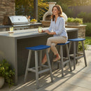 A woman sits on a Kanyon Living Poly Lumber Saddle Stool Bar Height Blue and Light Grey at a modern outdoor kitchen island, holding a drink and smiling. A grill, pizza oven, plants, and sunshine create a relaxed backyard atmosphere.
