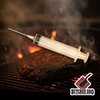 A Butcher BBQ Budget Meat Injector positioned over a piece of meat on a grill with flames in the background.