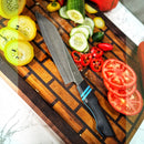 The Spaceman Knives Calypso Chef Knife, featuring a black and blue handle, rests on a wooden board amid sliced yellow and red tomatoes, cucumbers, and red chili peppers, ready for precision cutting.