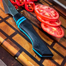A close-up of the Spaceman Knives Calypso Chef Knife with a black and blue handle on a wooden cutting board, demonstrating precision slicing beside tomatoes and chopped red chili peppers.