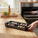 A person places a Napoleon Grills Cast Iron Jumbo Shrimp Tray (67734) with jumbo shrimp into an oven, while a wooden countertop, cutting board, bowl of lemons, and olive oil are visible in the background.
