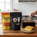 A hand stirs chili in a pot, seasoned with the Meat Church Chili & Cornbread Comfort Bundle. In front are two packages from Meat Church and a plate of cornbread, all set on a white countertop with a matching backsplash.