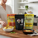 A woman prepares the Meat Church Chili & Cornbread Comfort Bundle in her kitchen, slicing cornbread on a wooden board with a skillet and whisk nearby, while packets of the bundle are displayed beside a bowl of chili.