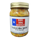 A glass jar labeled DDR BBQ Supply Crushed Olive Spread Pint – 16 oz shows a chopped olive mixture. The label features a red, white, and blue design with a white star emphasizing its Mediterranean flavor. Brand: DDR BBQ Supply.